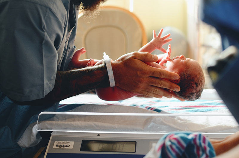 A male nurse in profile from the neck down. They’re holding a newborn baby over a scale