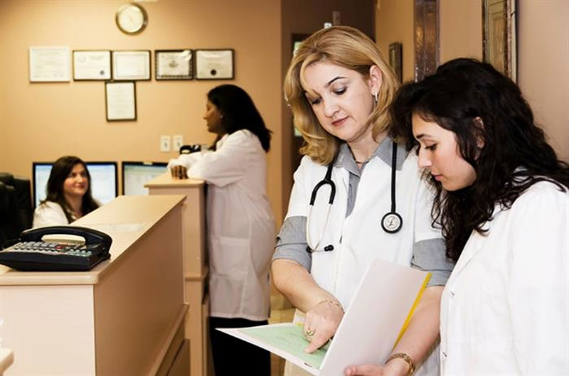 A group of four female doctors in a medical office. Two women are at the back chatting over a desk. Two women are at the front looking at a clipboard.